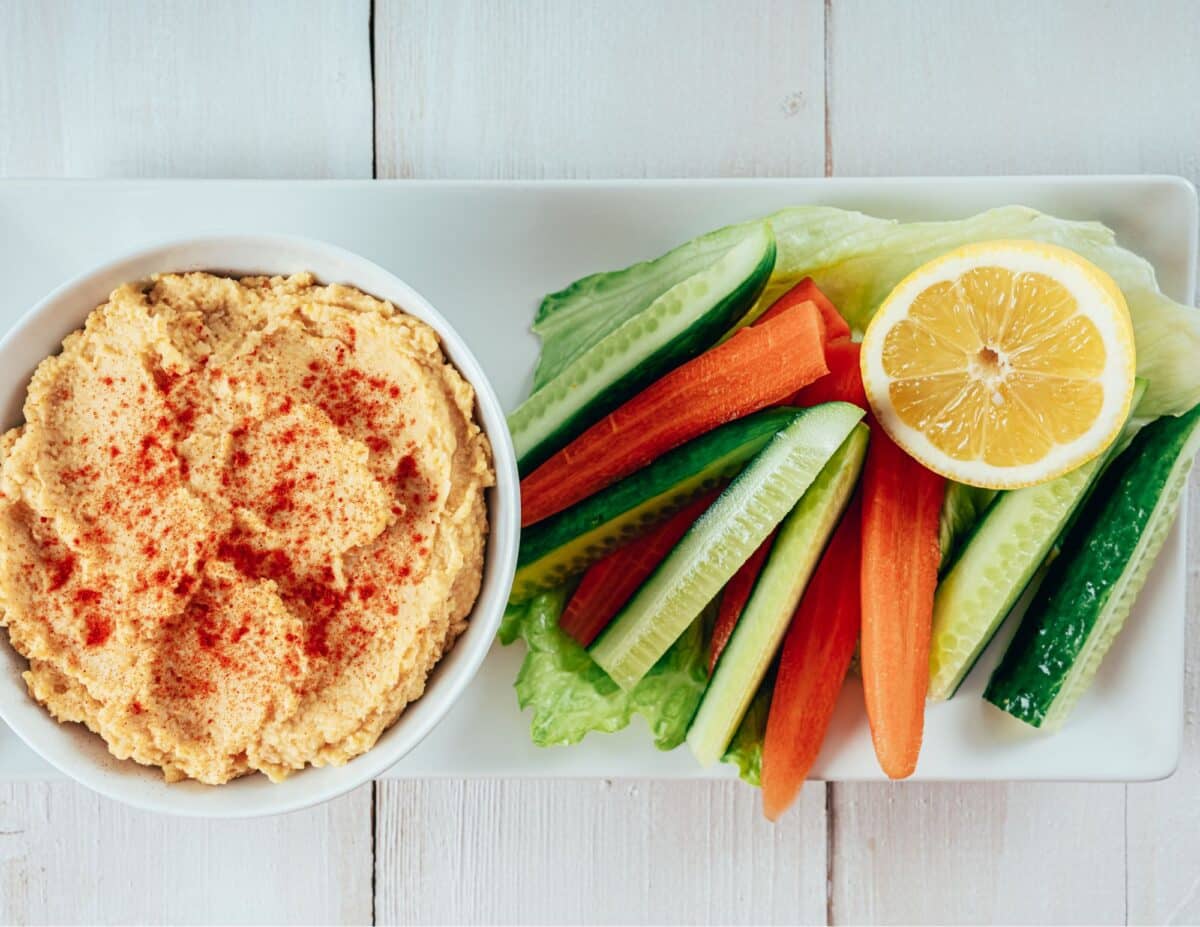 hummus in a bowl with another bowl filled up with cucumber and carrot sticks, garnished with a piece of lemon - cheap healthy snacks. 