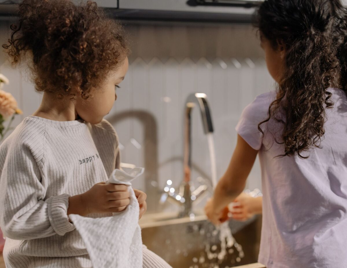 A girl is washing dishes while her younger sister is watching-chores for 8-year-olds.