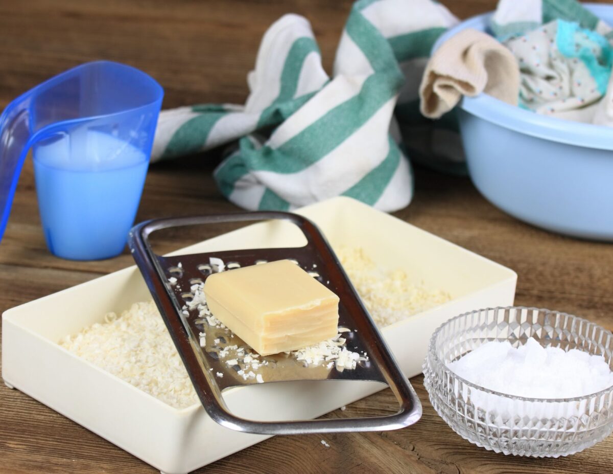 A container of washing soda, a bowl of baking soda and some soap being grated with a grater - DIY natural household cleaners.