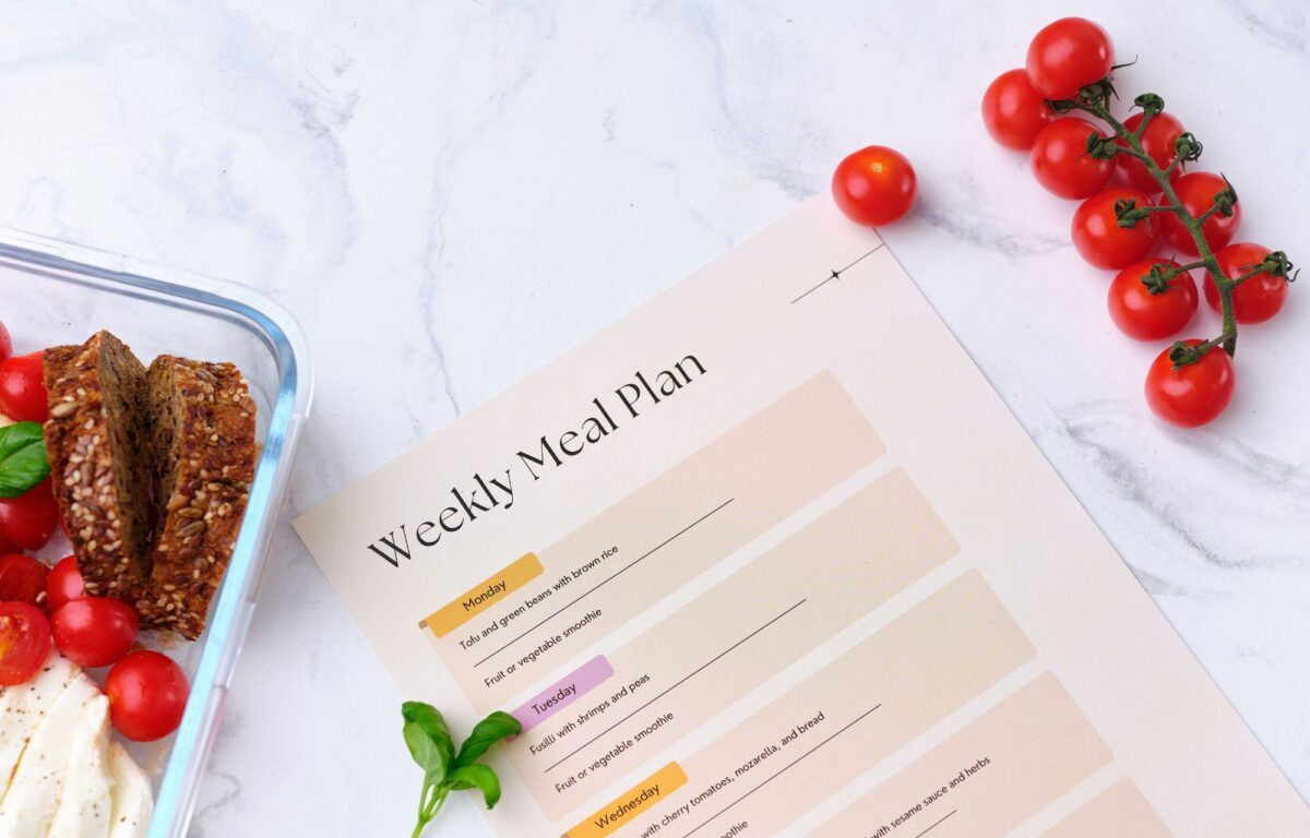 A document called, 'Weekly Meal Plan' is on a table with some food surrounding it - Frugalnook Category Meal Organization.