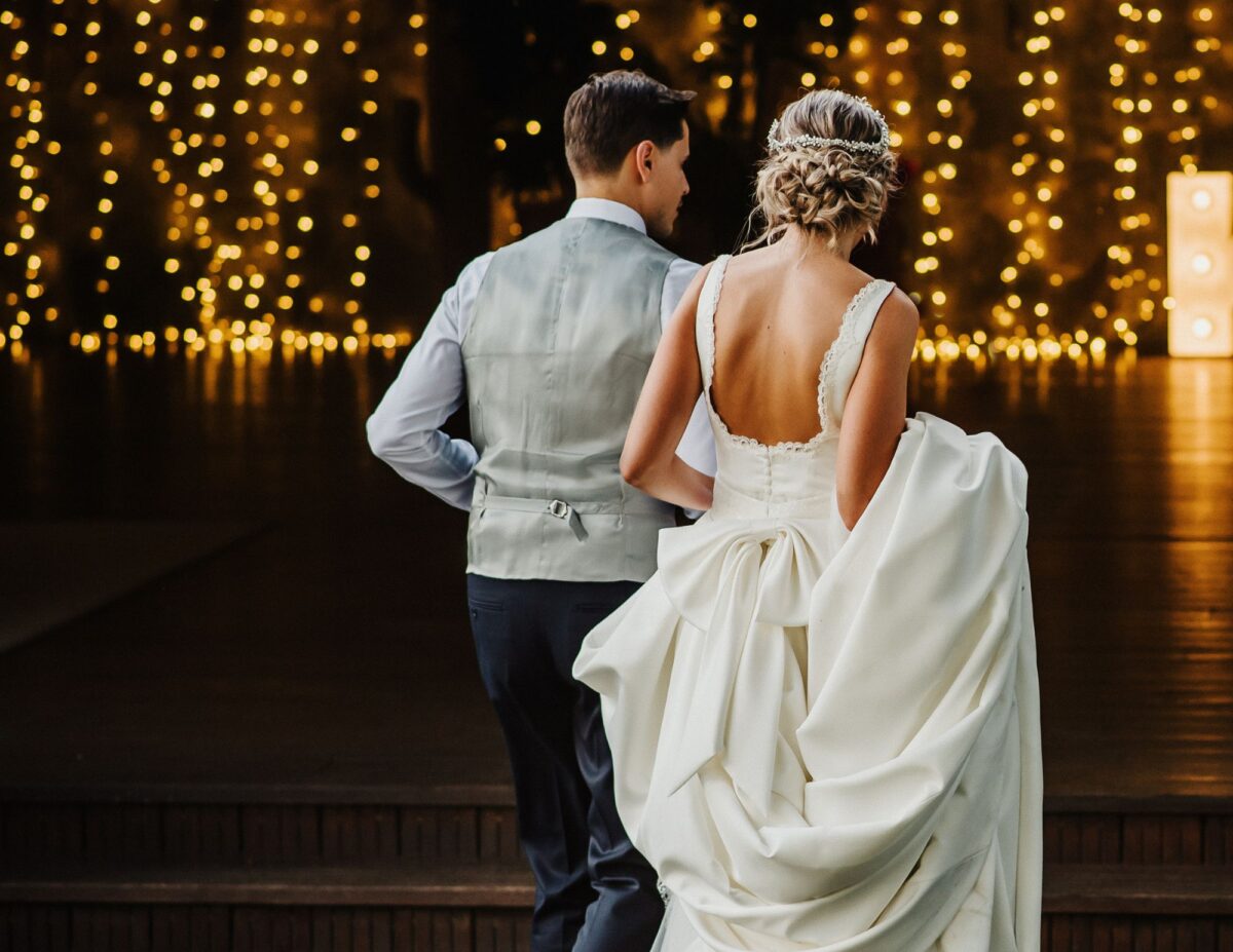 a bride and groom are walking near string lights - outdoor wedding ideas on a budget