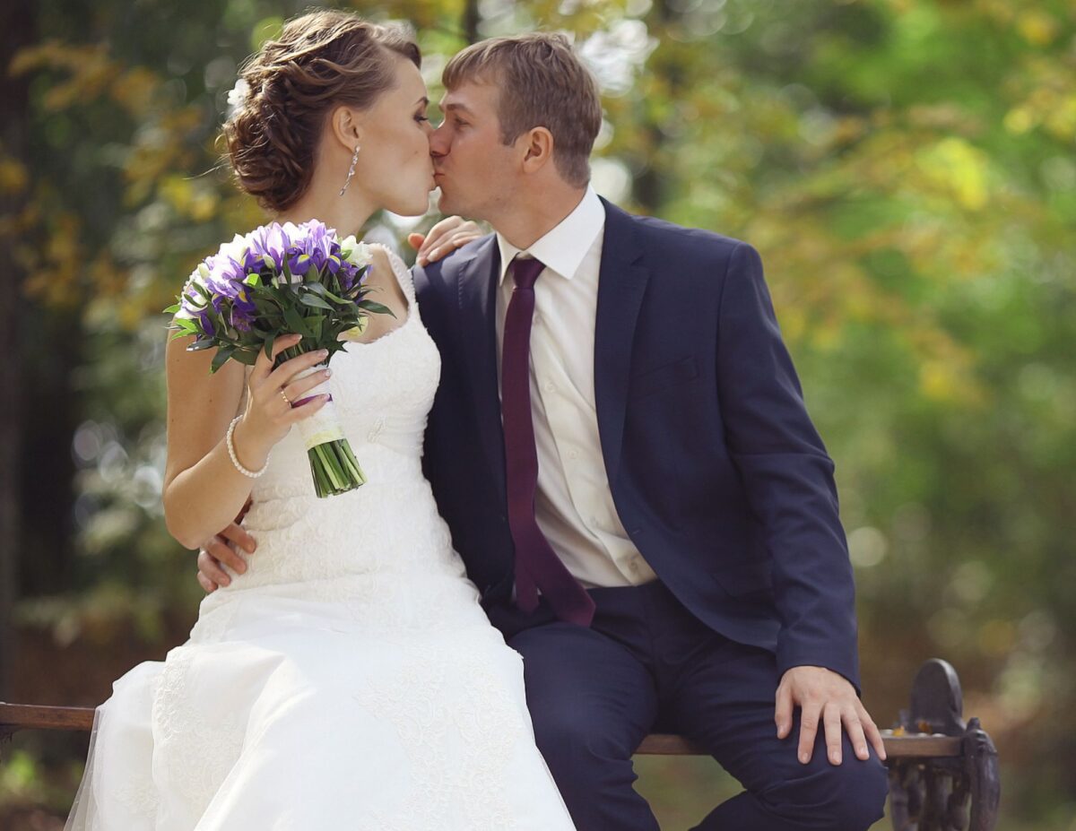a bride and groom are sitting on a park bench and kissing - outdoor wedding ideas on a budget