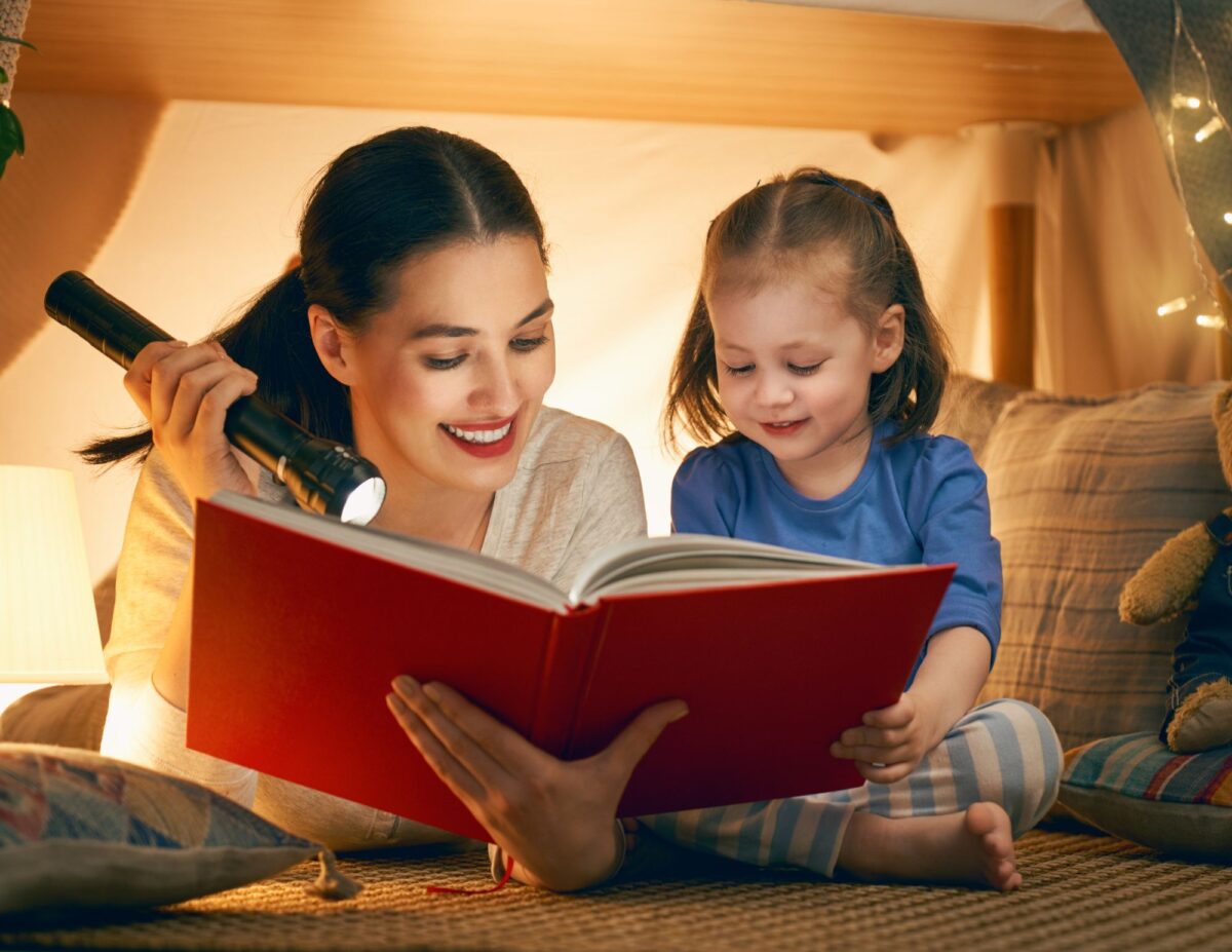 a woman is reading a book to a little girl by flashlight - benefits of being a stay-at-home mom