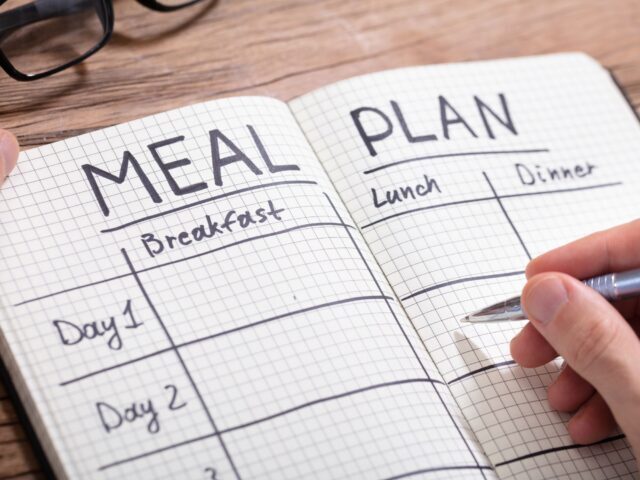 Someone is writing in a notebook that says, "Meal Plan: Breakfast, Lunch, Dinner" - how to save money on groceries without coupons
